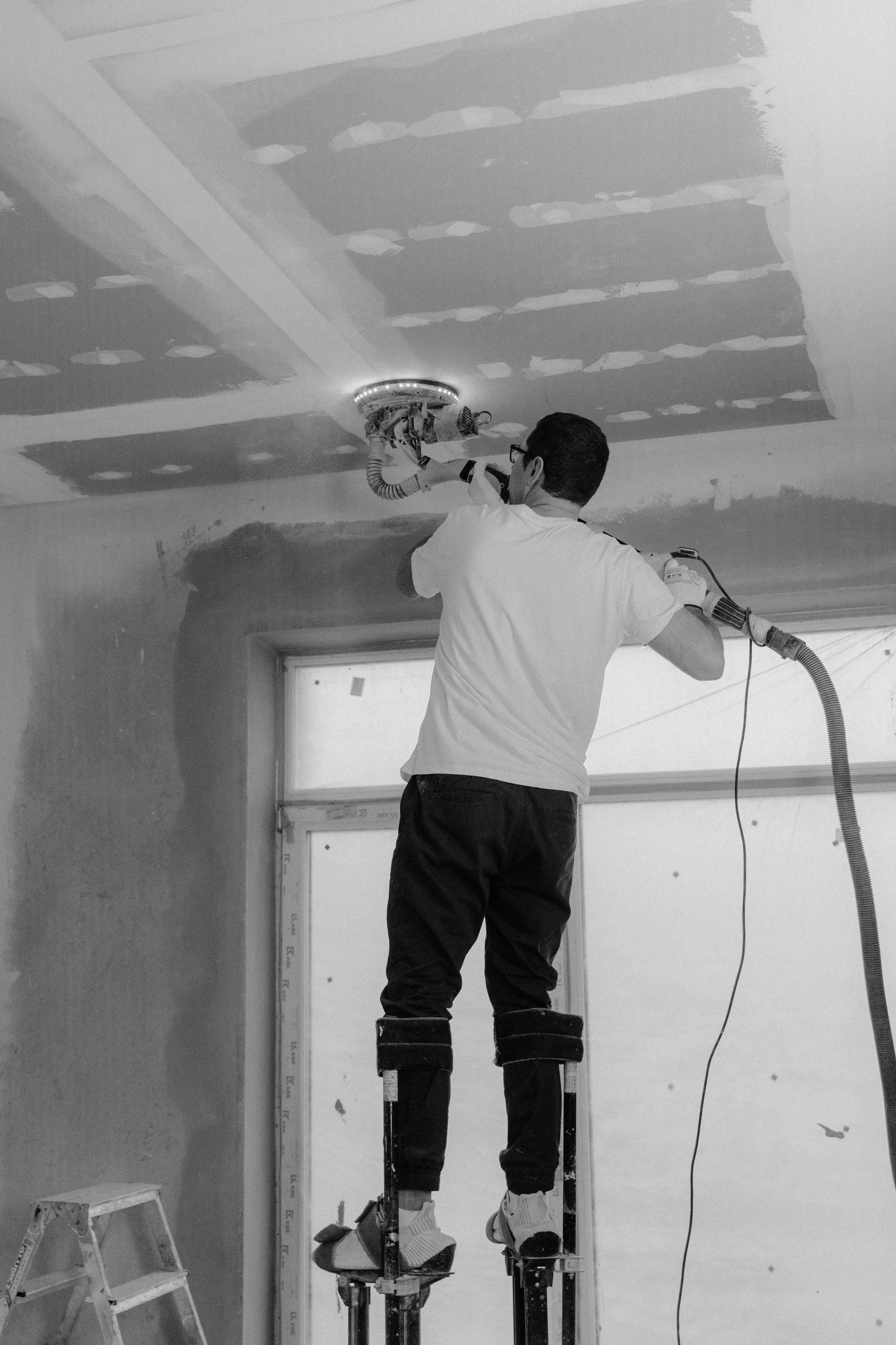 Man sanding ceiling drywall with a power sander while standing on stilts.