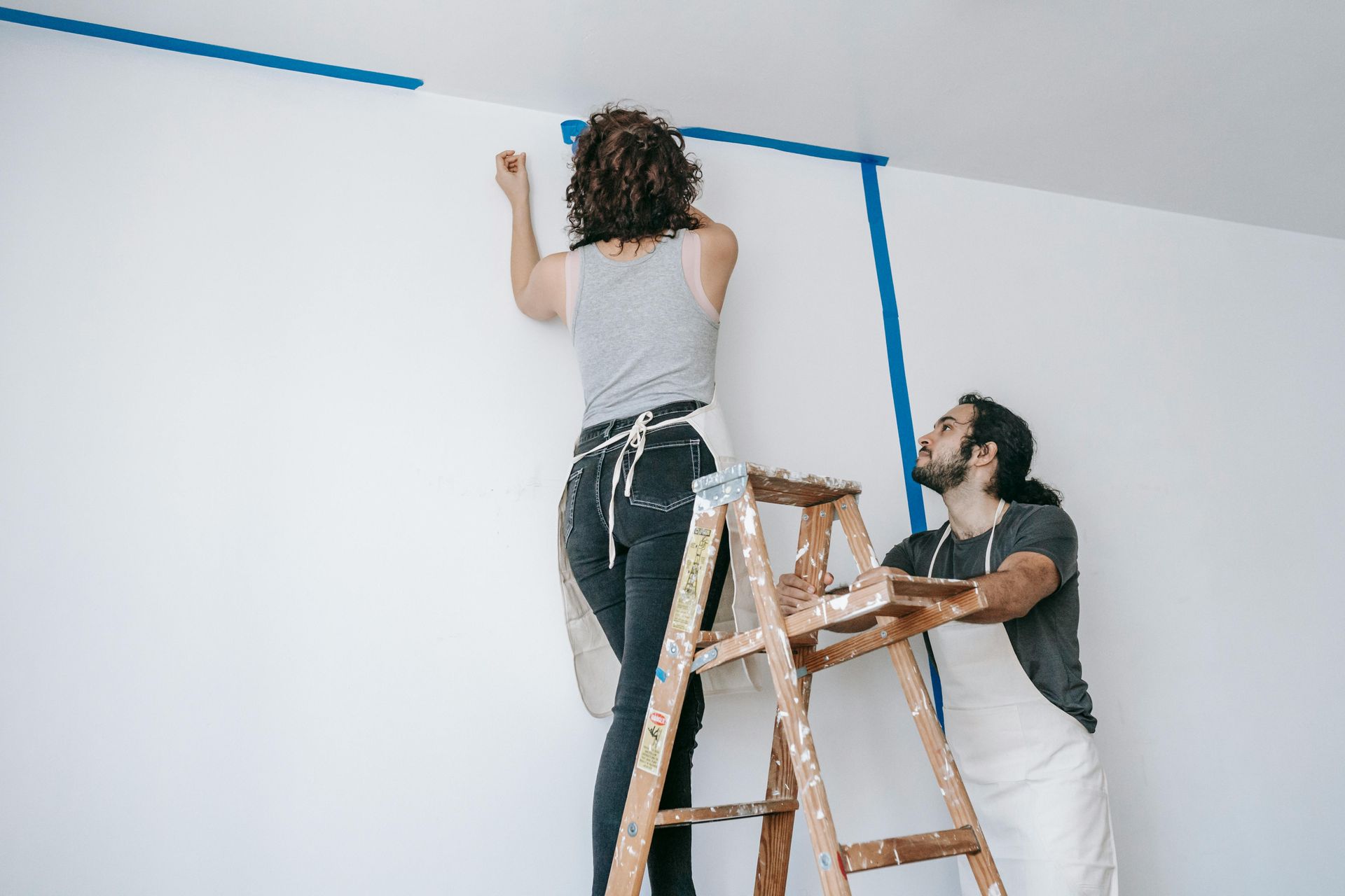 Woman on ladder taping wall, man assisting. White wall, blue tape, aprons.