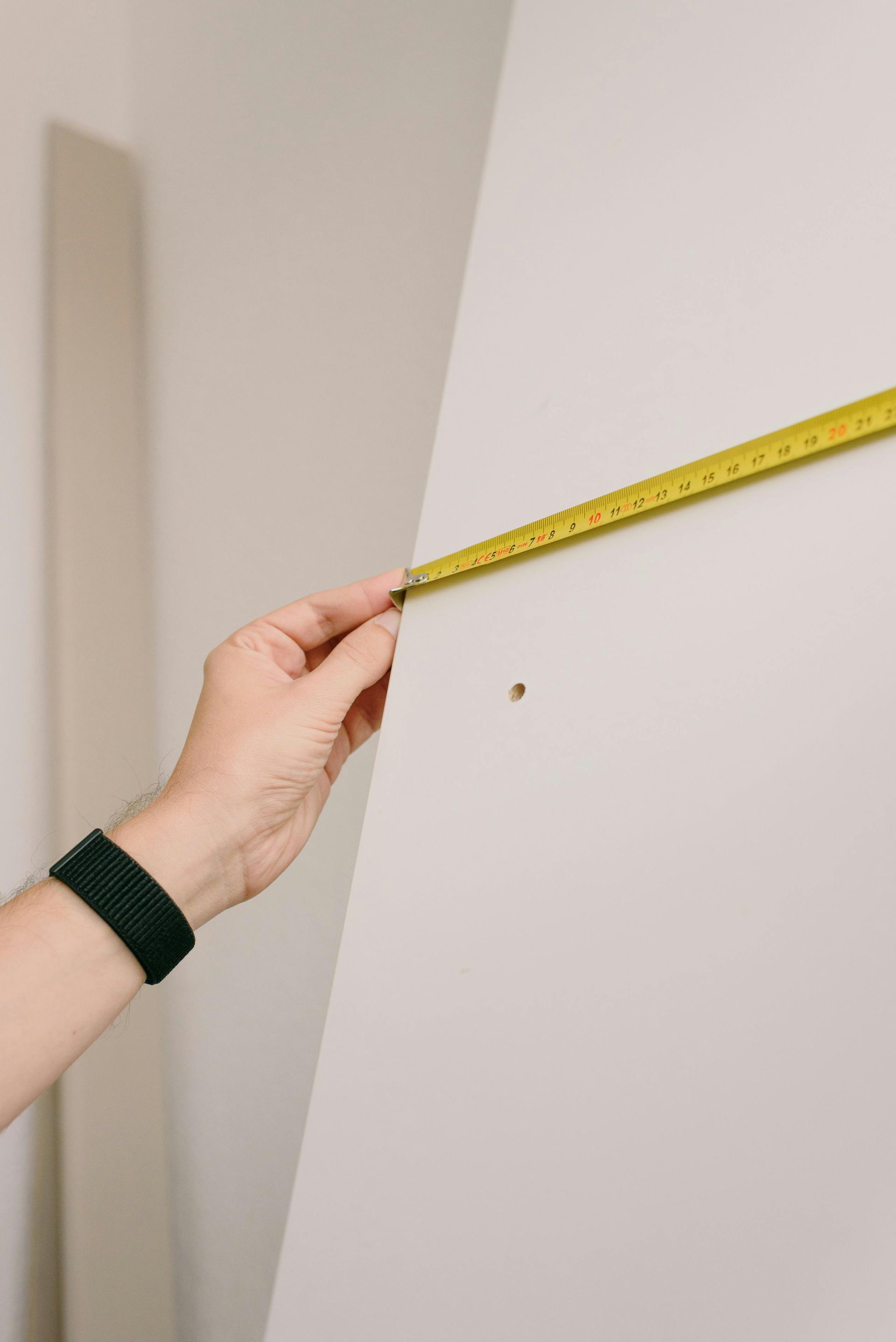 Hand holding a yellow tape measure against a white surface, measuring a point near a small hole.