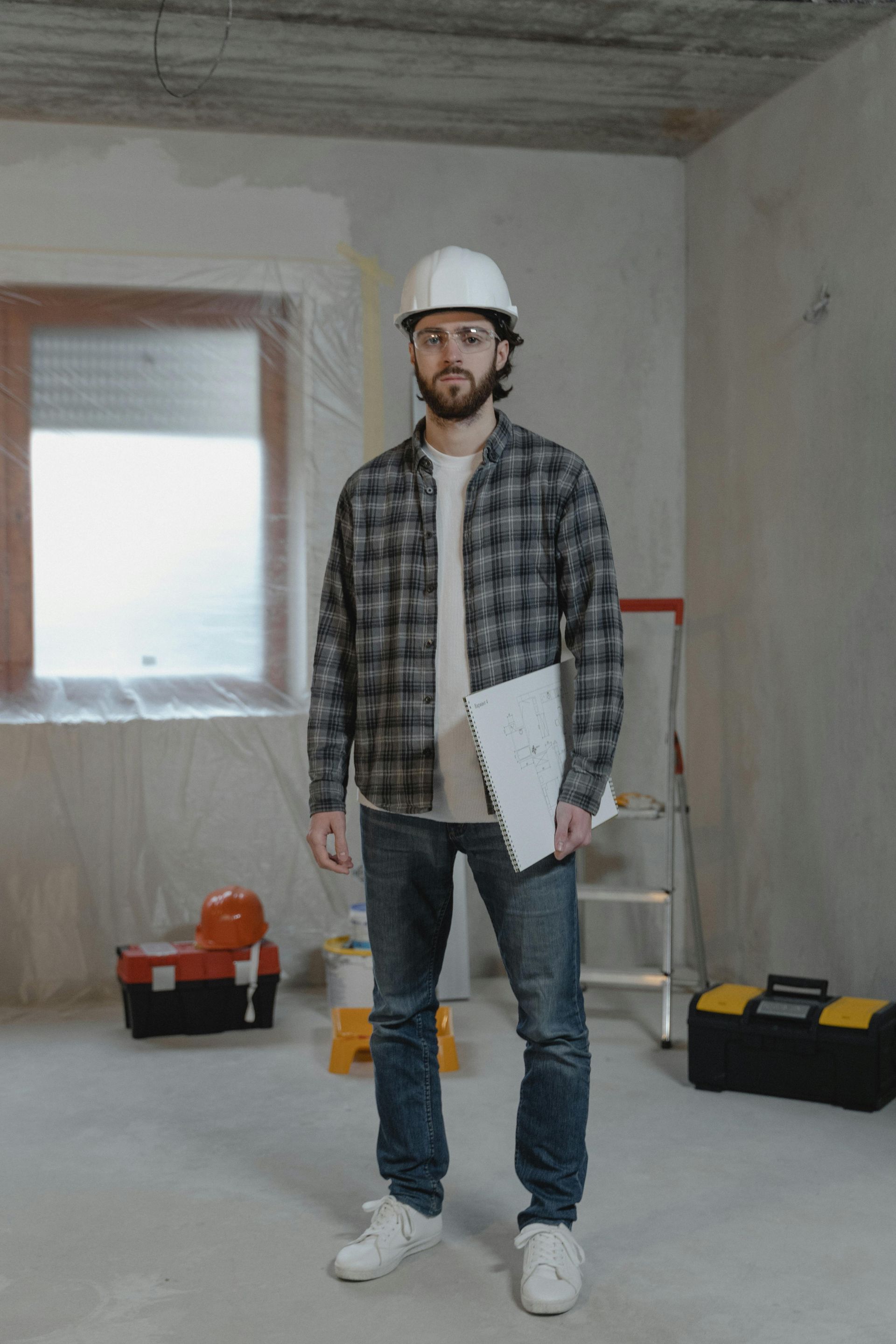 Construction worker in hard hat, plaid shirt, and jeans, holding a clipboard in a construction zone.