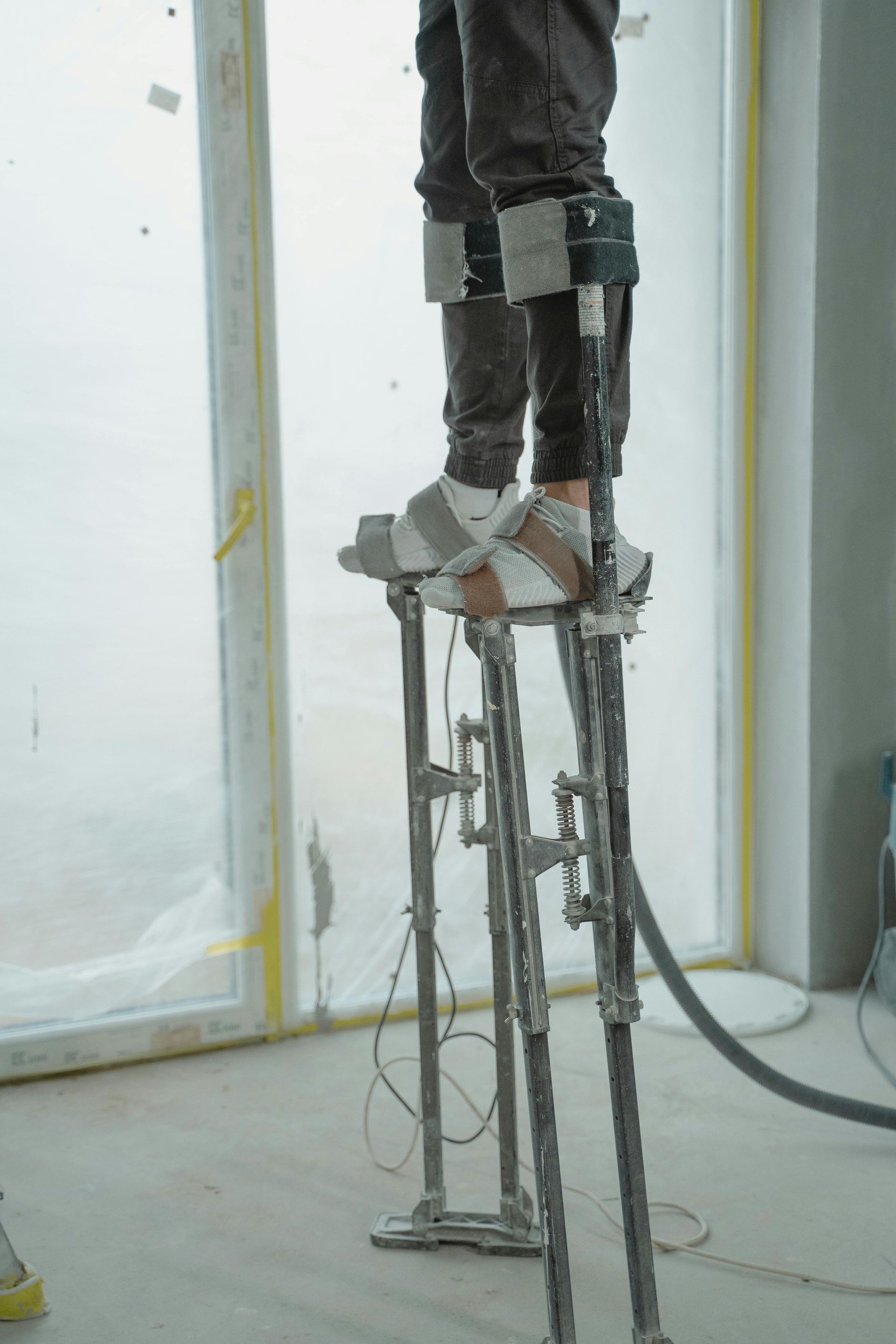 Person wearing adjustable stilts, standing near a window. Gray metal stilts, gray pants, interior setting.