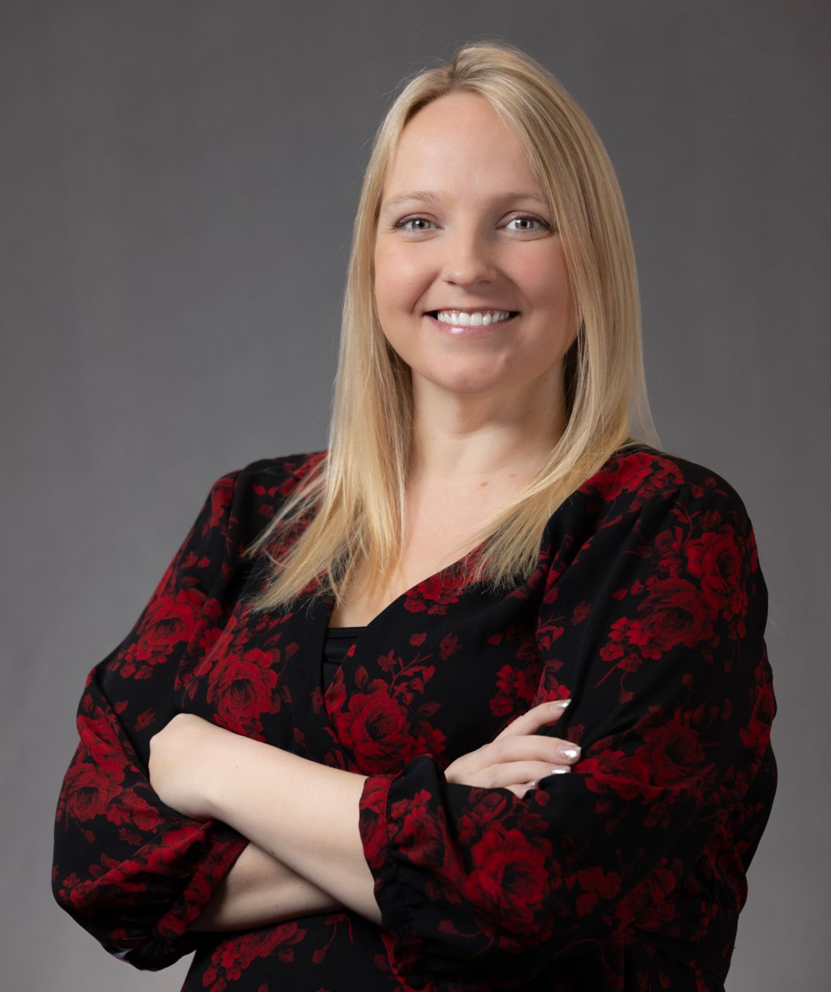 A woman in a red and black floral shirt is smiling with her arms crossed.