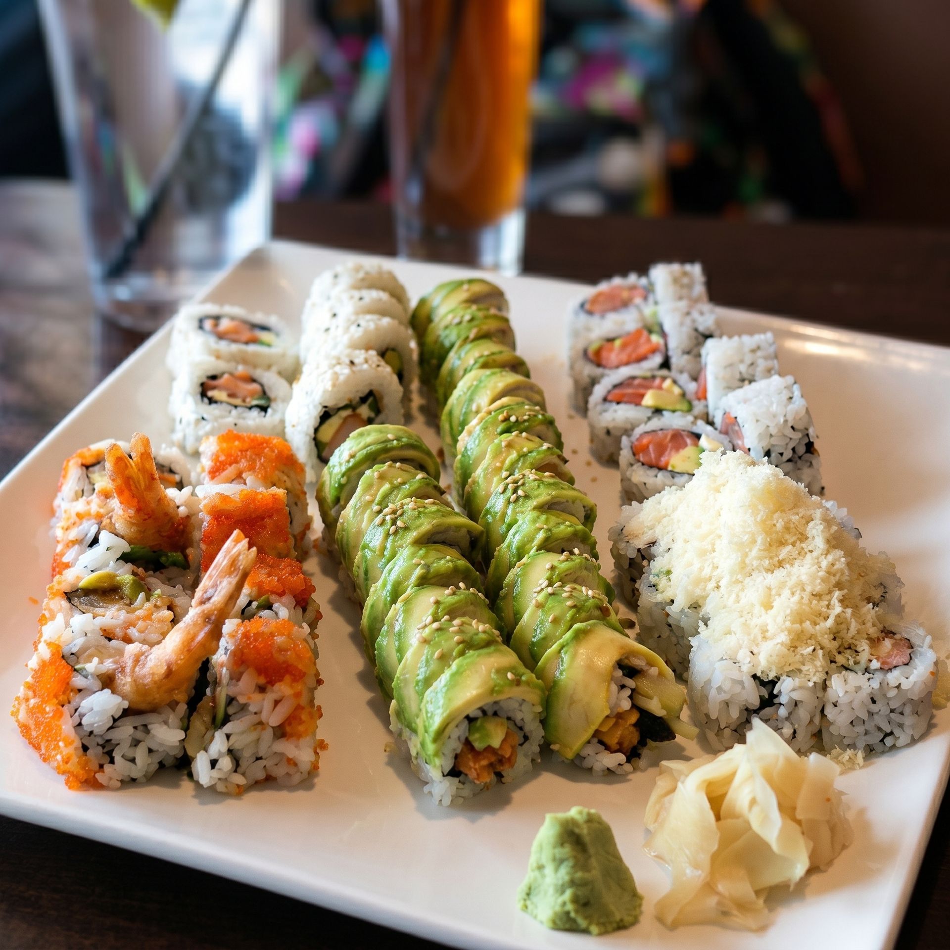 Assorted sushi rolls on a white plate, topped with avocado, fish roe, and sesame seeds, with wasabi and ginger.