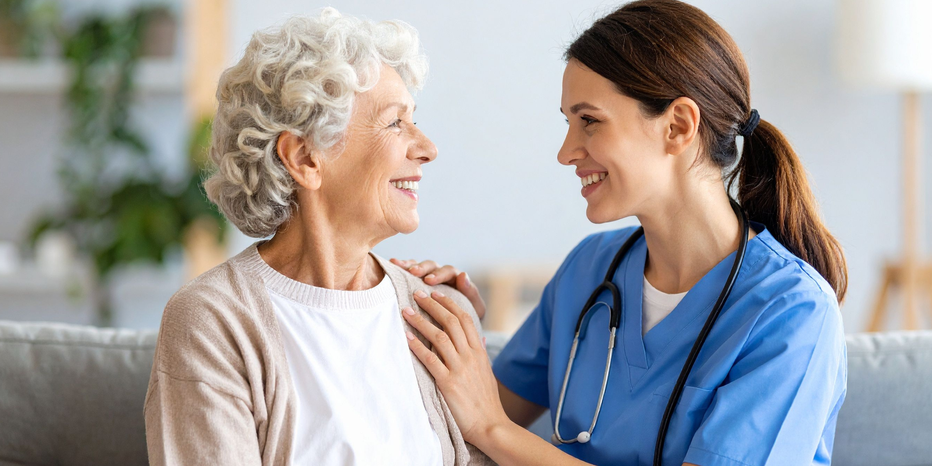 A smiling medical professional in scrubs comforts an older woman on a couch.