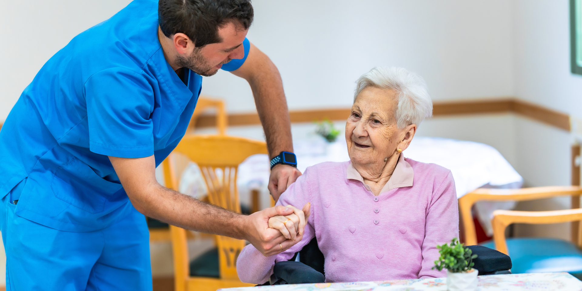 A healthcare worker in blue scrubs assisting an elderly person sitting at a table in a care facility.
