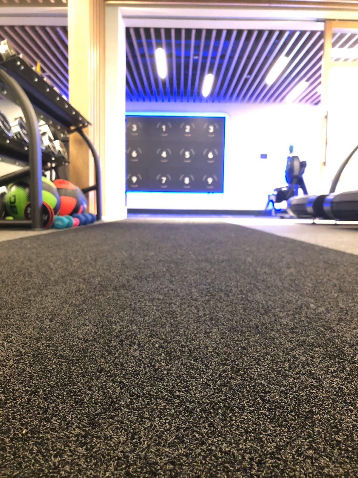 Black textured gym floor leading towards a training area with equipment and a person walking in the distance.