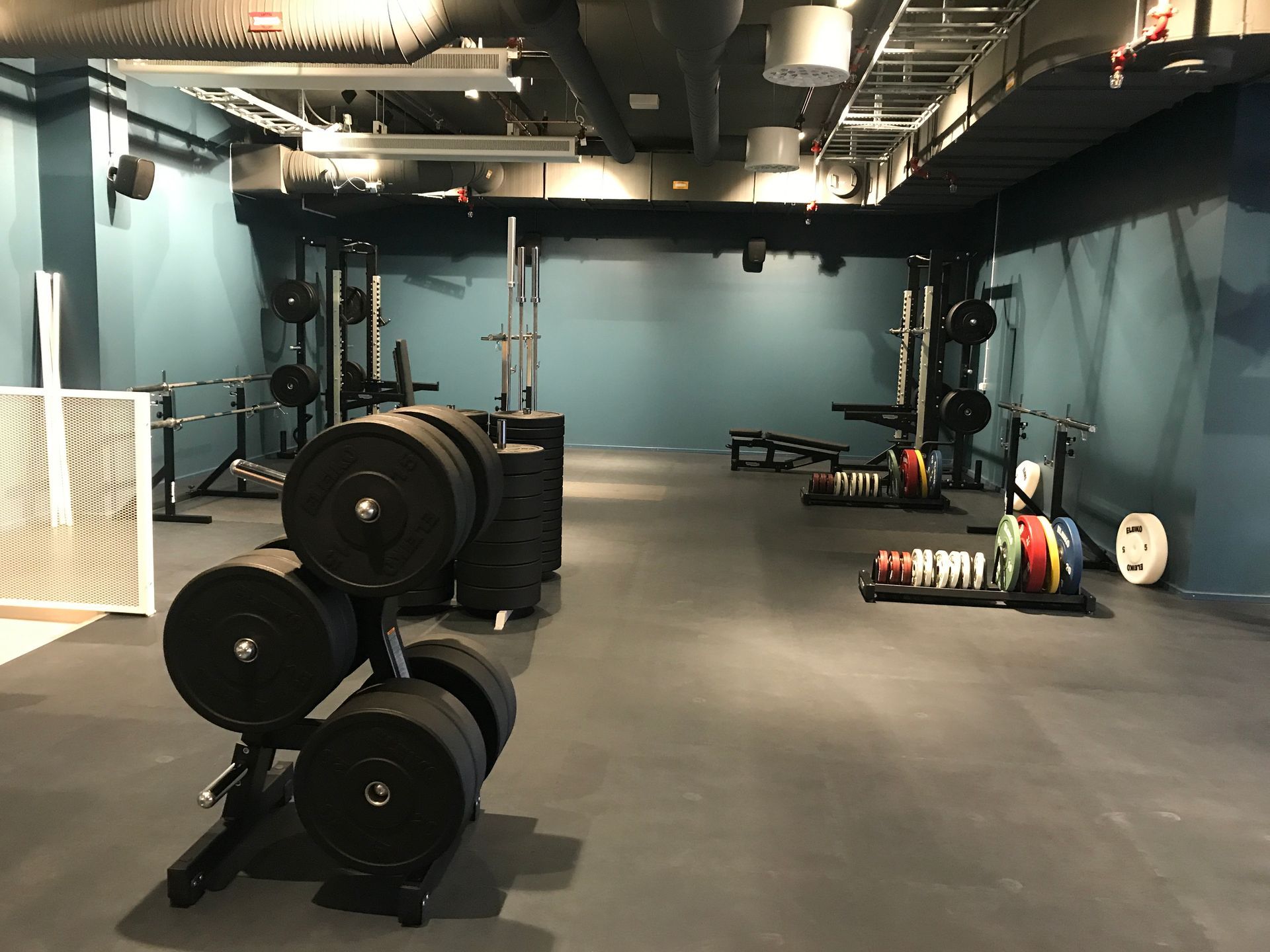 Weightlifting gym with weight racks, barbells, and weights. Dark gray floor, blue walls, and exposed ceiling fixtures.