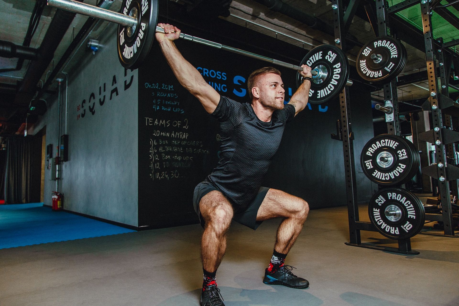 Man in gym performs overhead squat with barbell, wearing black athletic wear.