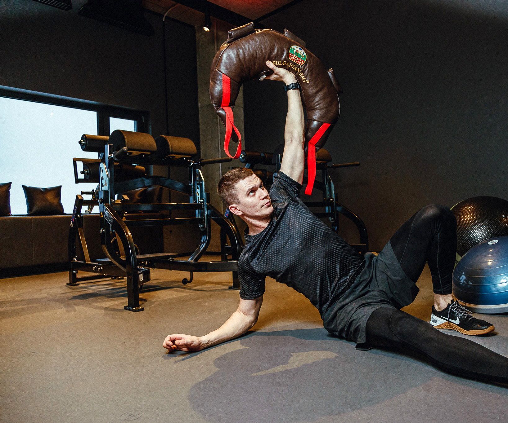 Man exercising with a sandbag in a gym, lying on his side, lifting overhead.