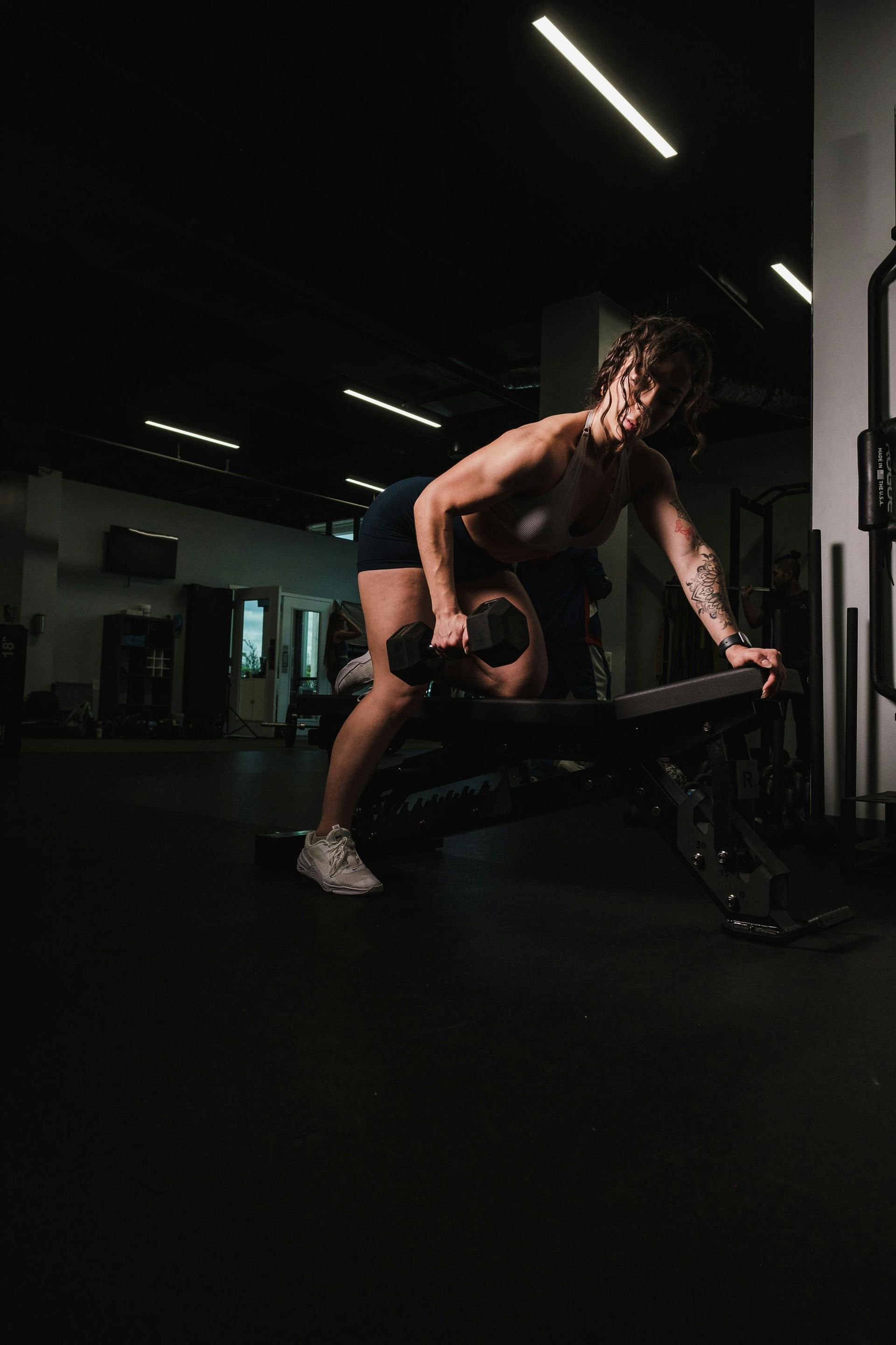 Woman doing dumbbell rows on a weight bench in a gym. She is leaning forward with one arm extended, dark setting.