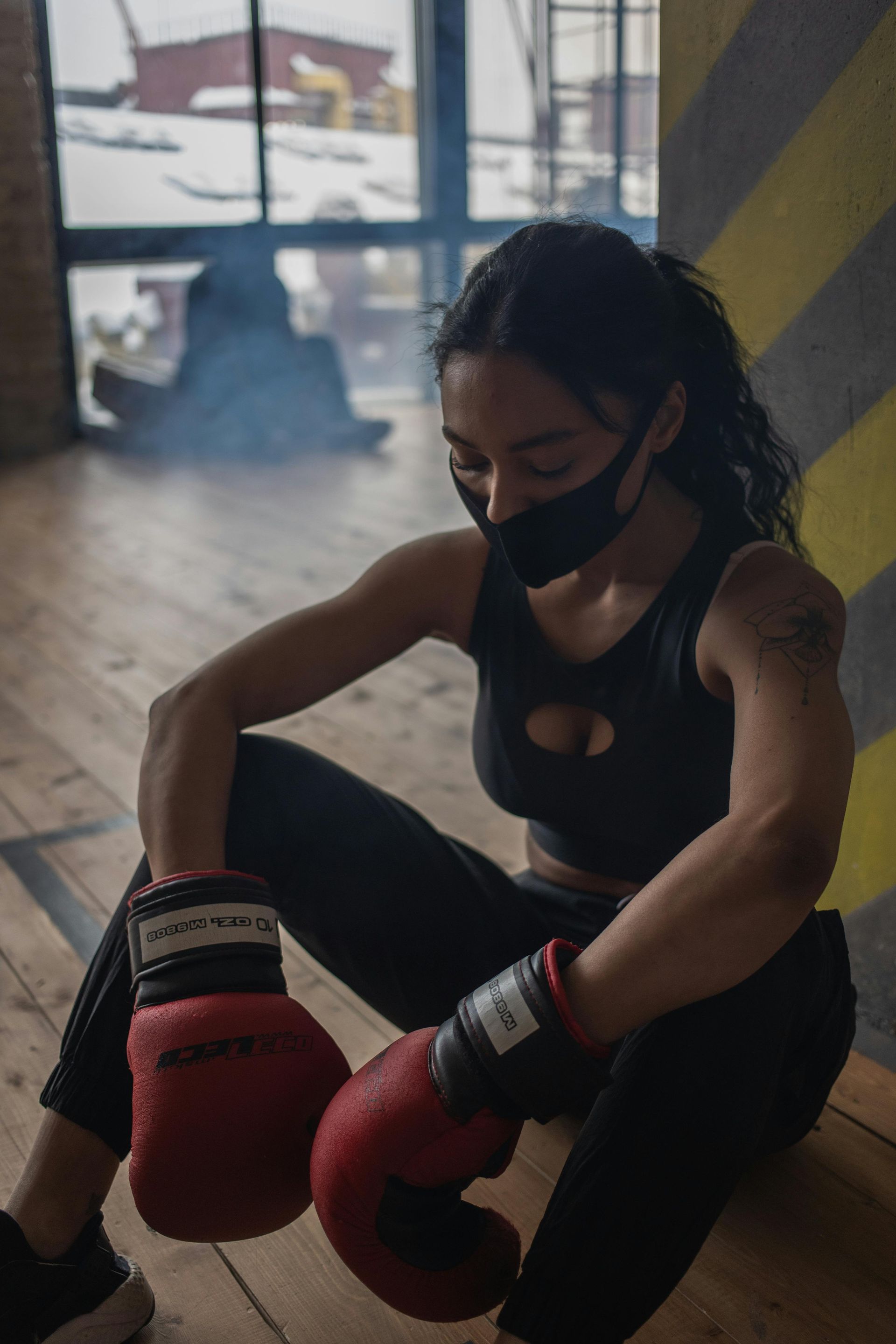 Woman in black athletic wear and a face mask rests with boxing gloves in a gym.