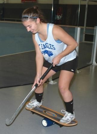 A girl wearing a eagles shirt is balancing a hockey stick on a skateboard