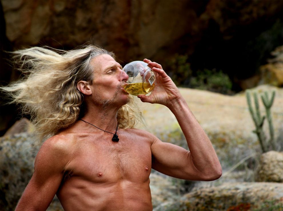 A shirtless man with long hair is drinking from a bottle.