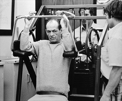 A man is using a machine in a gym while another man watches