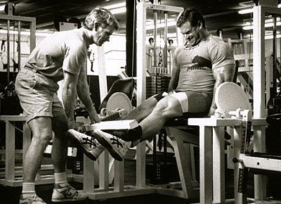 A man is helping another man do leg exercises in a gym.