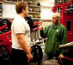 A man in a green jacket is talking to a man in a white shirt in a gym.