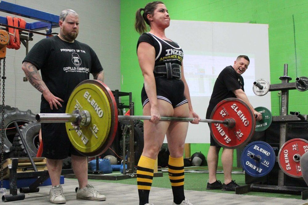 A woman is lifting a barbell in a gym while two men watch.