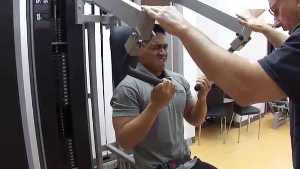 A man is using a machine in a gym while another man watches.