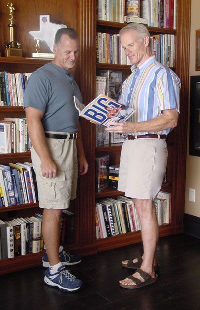 Two men are standing in front of a bookshelf looking at a book titled big