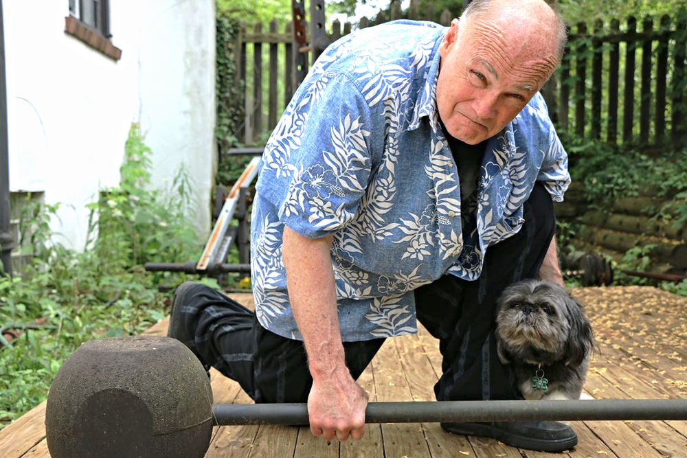 A man is kneeling down next to a small dog while holding a barbell.