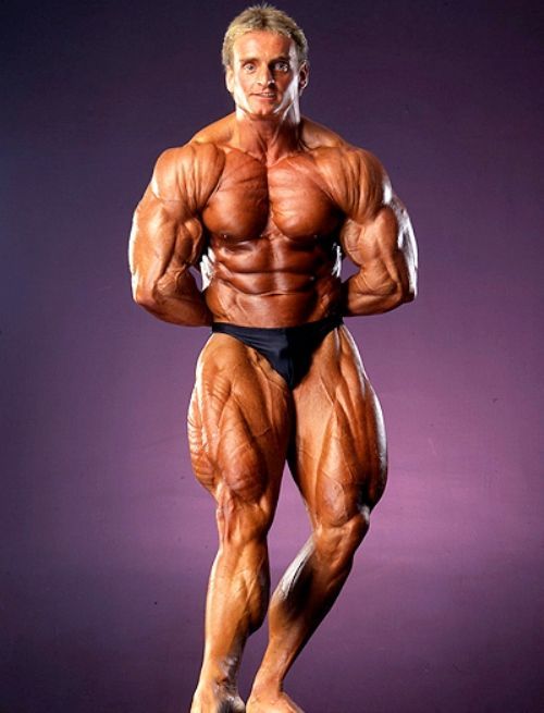 A bodybuilder is standing with his arms crossed in front of a purple background