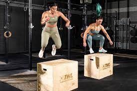 Two women are jumping over wooden boxes in a gym.