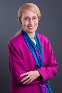 A woman in a purple jacket and scarf is smiling with her arms crossed.