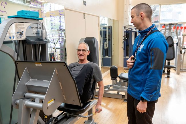 A man is sitting on a leg press machine in a gym while a man talks to him.