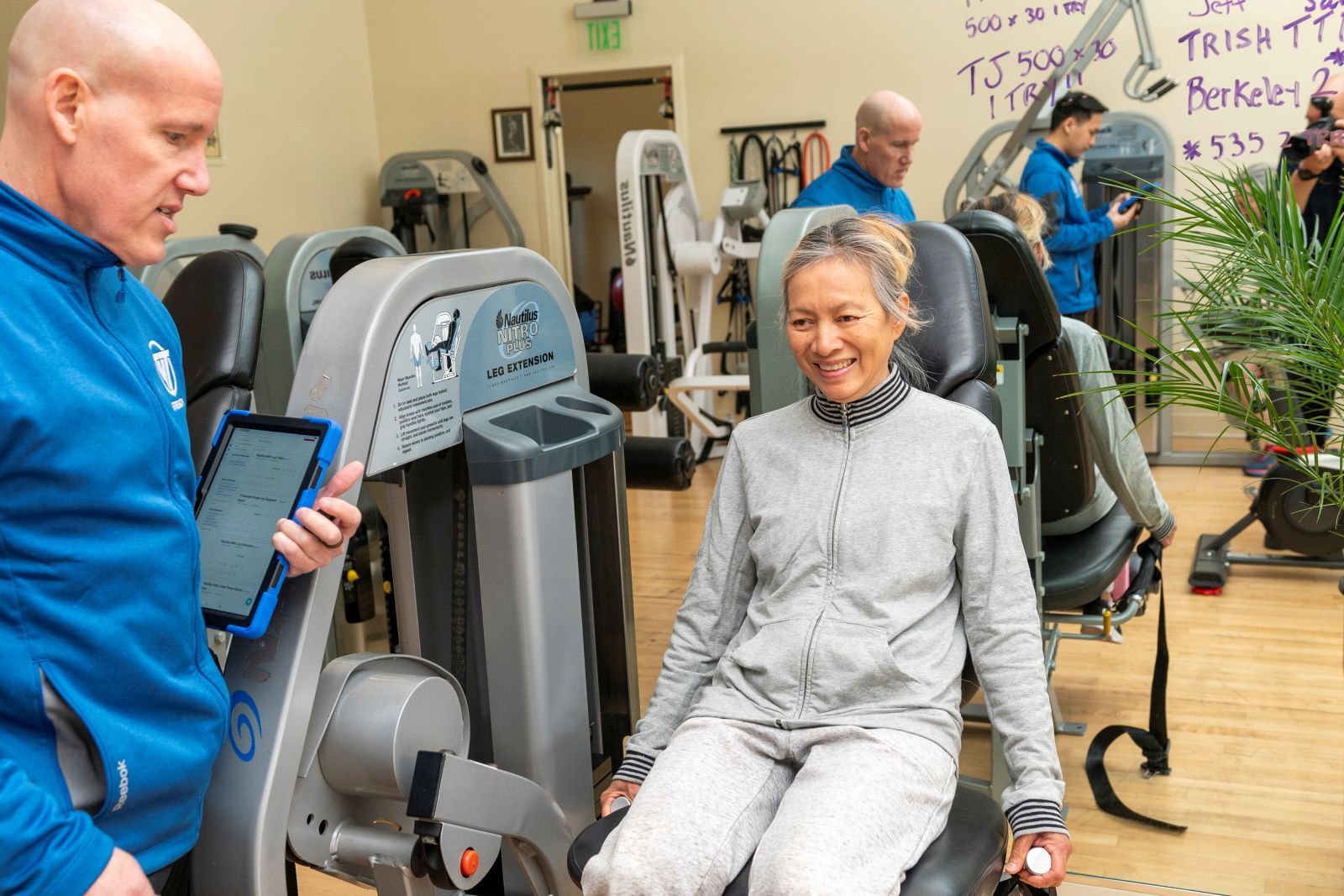 A man is talking to a woman sitting on a machine in a gym.