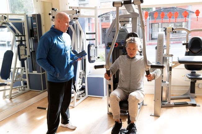 A man is standing next to a woman sitting on a machine in a gym.