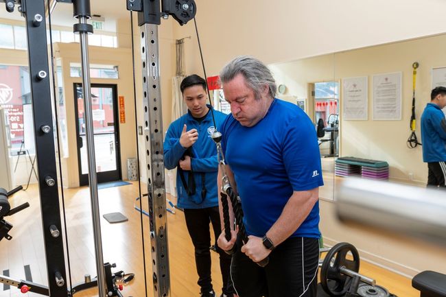 A man is using a machine in a gym while another man watches.