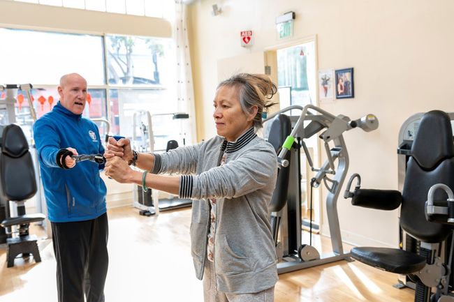 A man is helping an older woman do exercises in a gym.