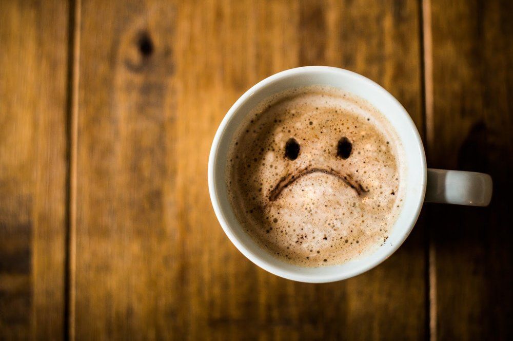 A cup of coffee with a sad face drawn on it on a wooden table.