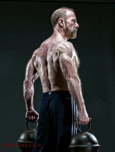 A shirtless man with a beard is holding two dumbbells behind his back.