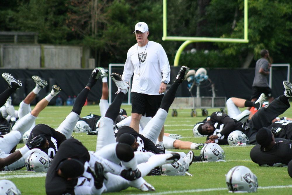 A group of football players are stretching on the field while a coach watches.