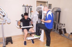 A man is sitting on a machine in a gym while another man looks on.