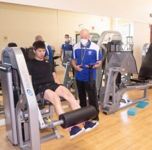 A man wearing a mask is standing next to a man sitting on a machine in a gym.