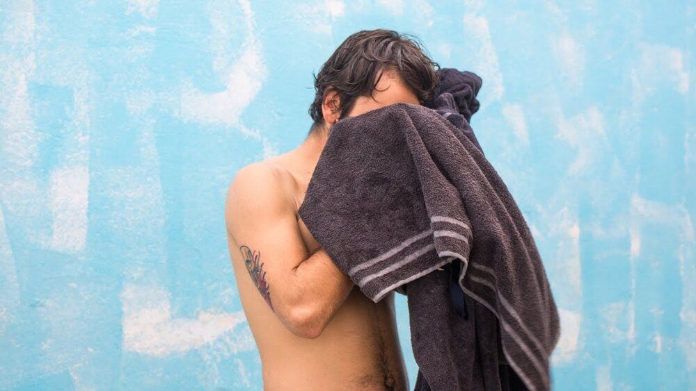 A shirtless man is covering his face with a towel.