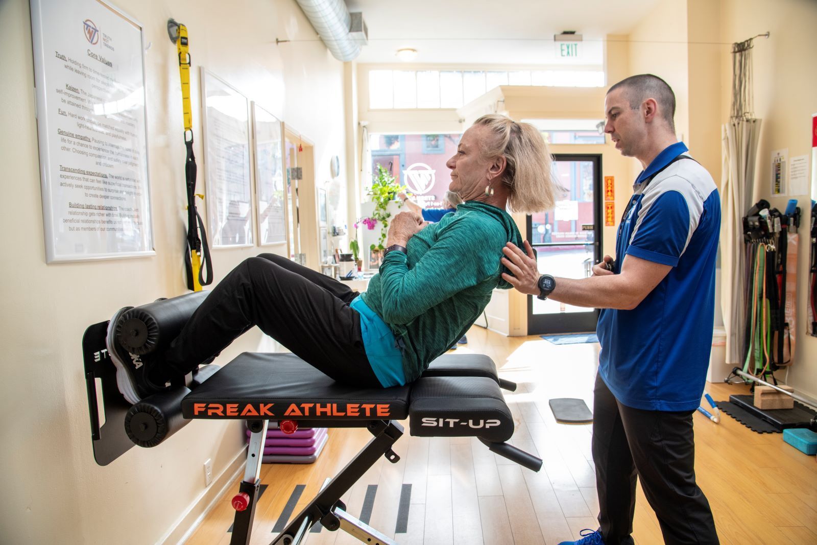 A man is helping a woman do exercises on a bench in a gym.