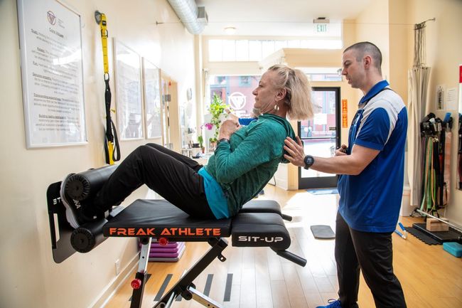 A man is helping a woman do exercises on a bench in a gym.