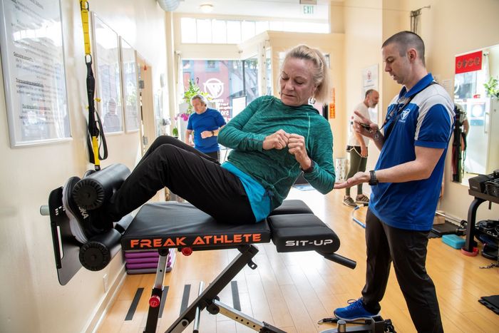 A man is helping a woman do exercises on a bench in a gym.