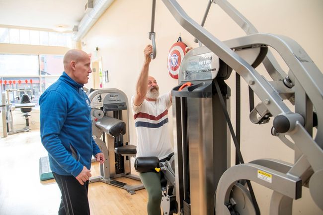 A man is helping another man use a machine in a gym.