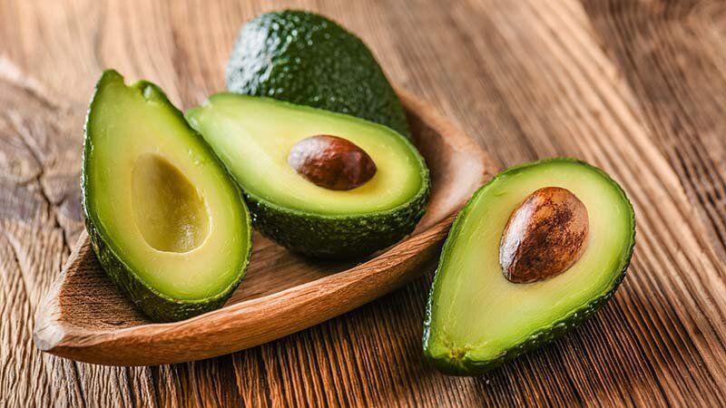 Three avocados are sitting on a wooden plate on a wooden table.