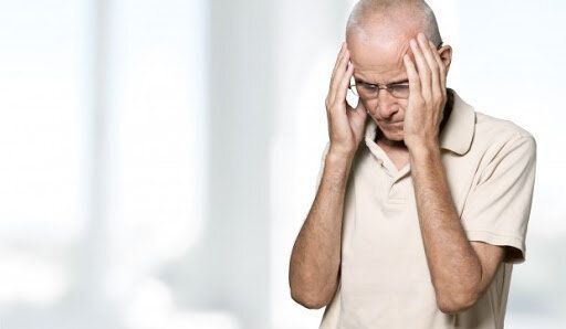 An older man is holding his head in pain while talking on a cell phone.