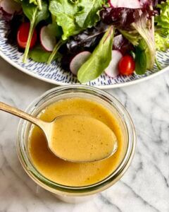 A bowl of salad dressing with a spoon in it next to a plate of salad.