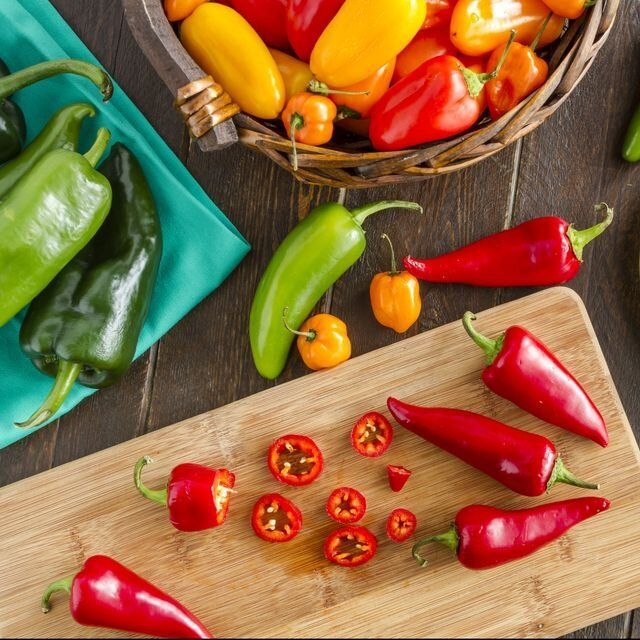 A wooden cutting board surrounded by peppers and a basket of peppers