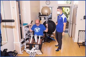 A man is using a machine in a gym while another man watches.