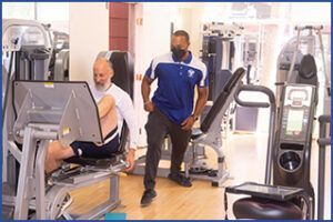 A man is standing next to a man sitting on a machine in a gym.