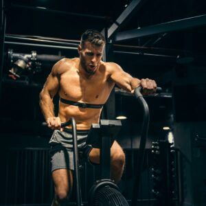 A shirtless man is riding an exercise bike in a gym.