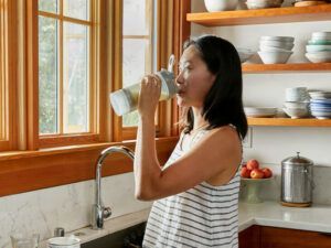 A woman is drinking water from a bottle in a kitchen.